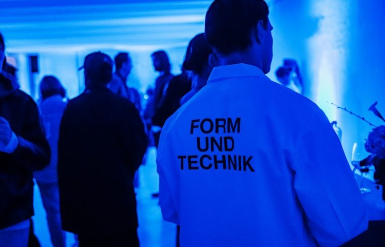 A man wearing a T-shirt with the words “Form und Technik” in a project room illuminated by blue light.