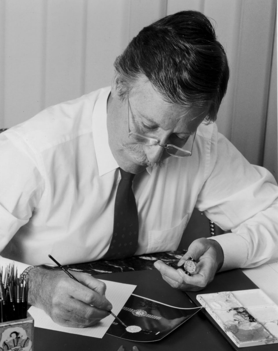 Photo noir et blanc du légendaire créateur de montres IWC Gérald Genta en chemise blanche dessinant à son bureau.