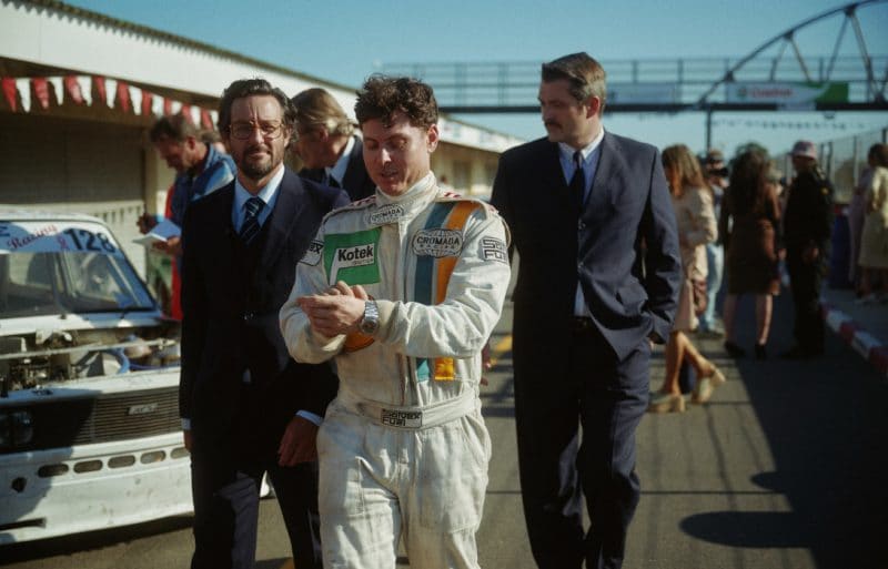 Racing driver with uniform wearing the IWC Ingenieur SL with two men in suits in background.,Racing driver with uniform wearing the IWC Ingenieur SL with two men in suits in background.