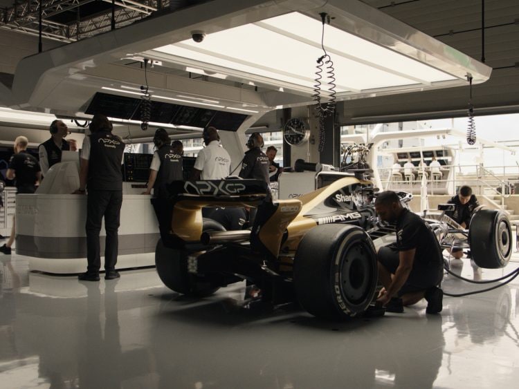 Mechanics work on a Formula 1 car in a modern garage, with bright overhead lights and equipment in the background.